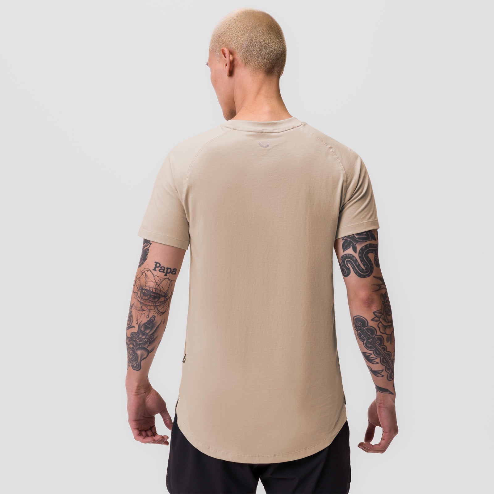 Beige Supima® Established Tee worn by model, back view, showing curved hem and sleeve tattoos