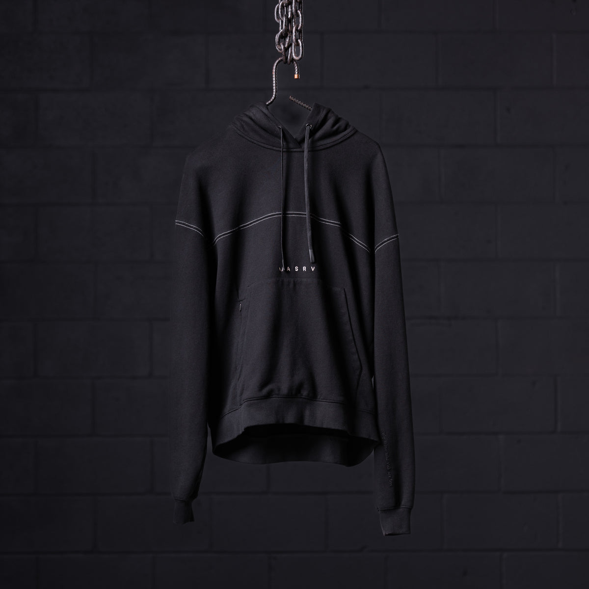 Black contrast thread hoodie displayed on hanger against dark brick wall