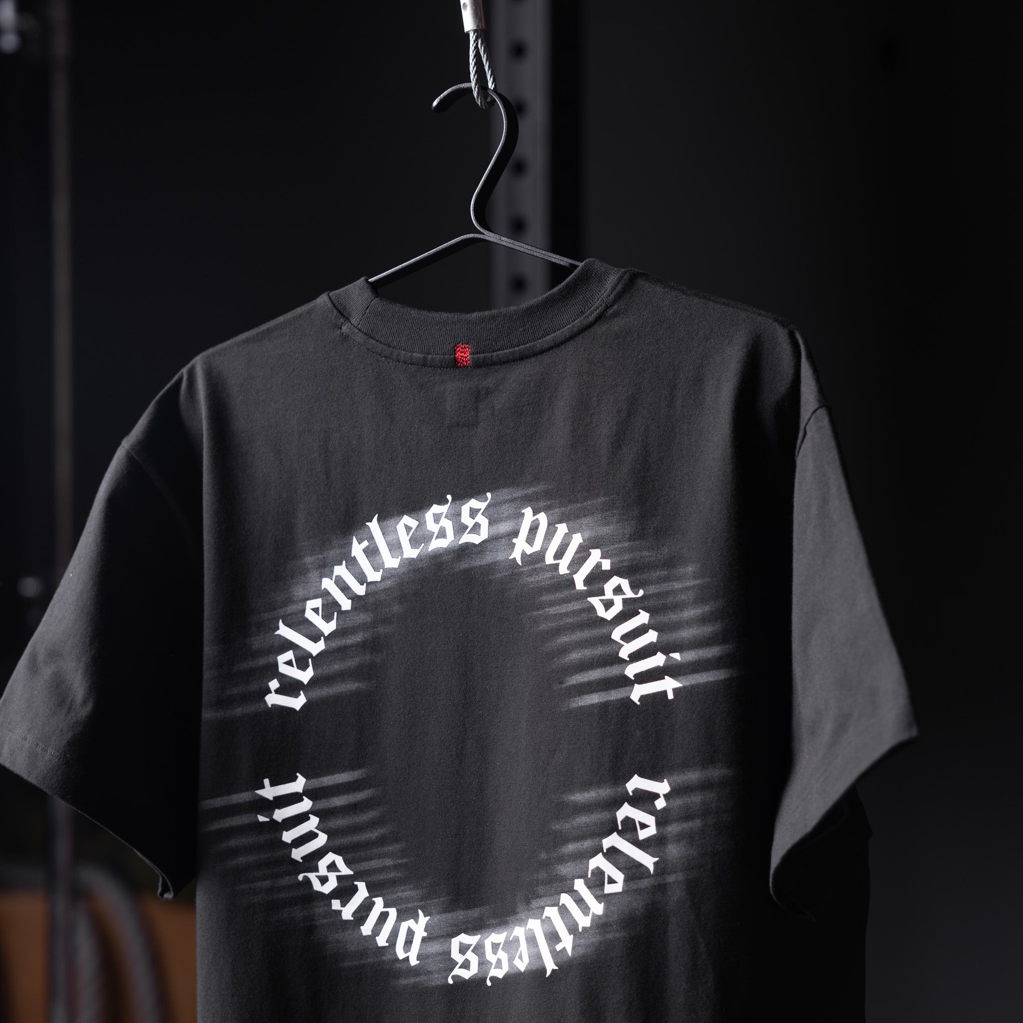 Black Tech Essential Relaxed Tee with 'relentless pursuit' circular text design on hanger