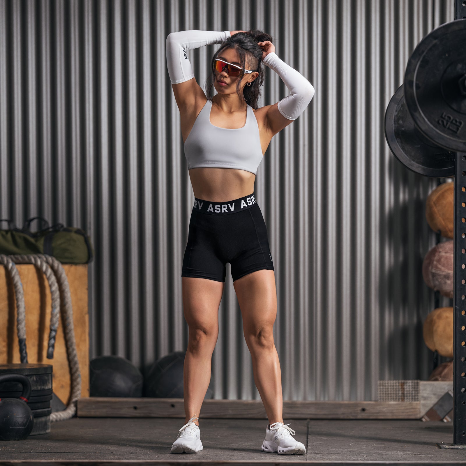 Black Lycra ribbed shorts worn with white sports bra and arm sleeves, model posing against corrugated metal backdrop
