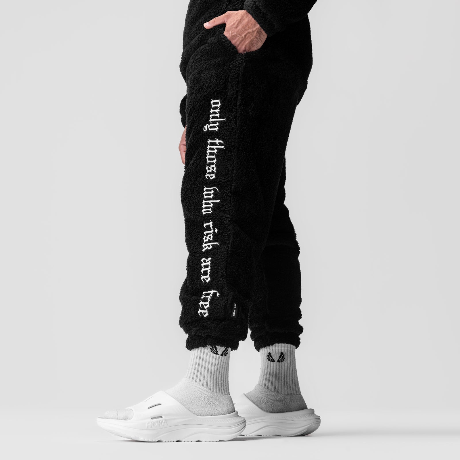 Black sherpa recovery sweats with white text on model wearing white socks and slides, lower body view