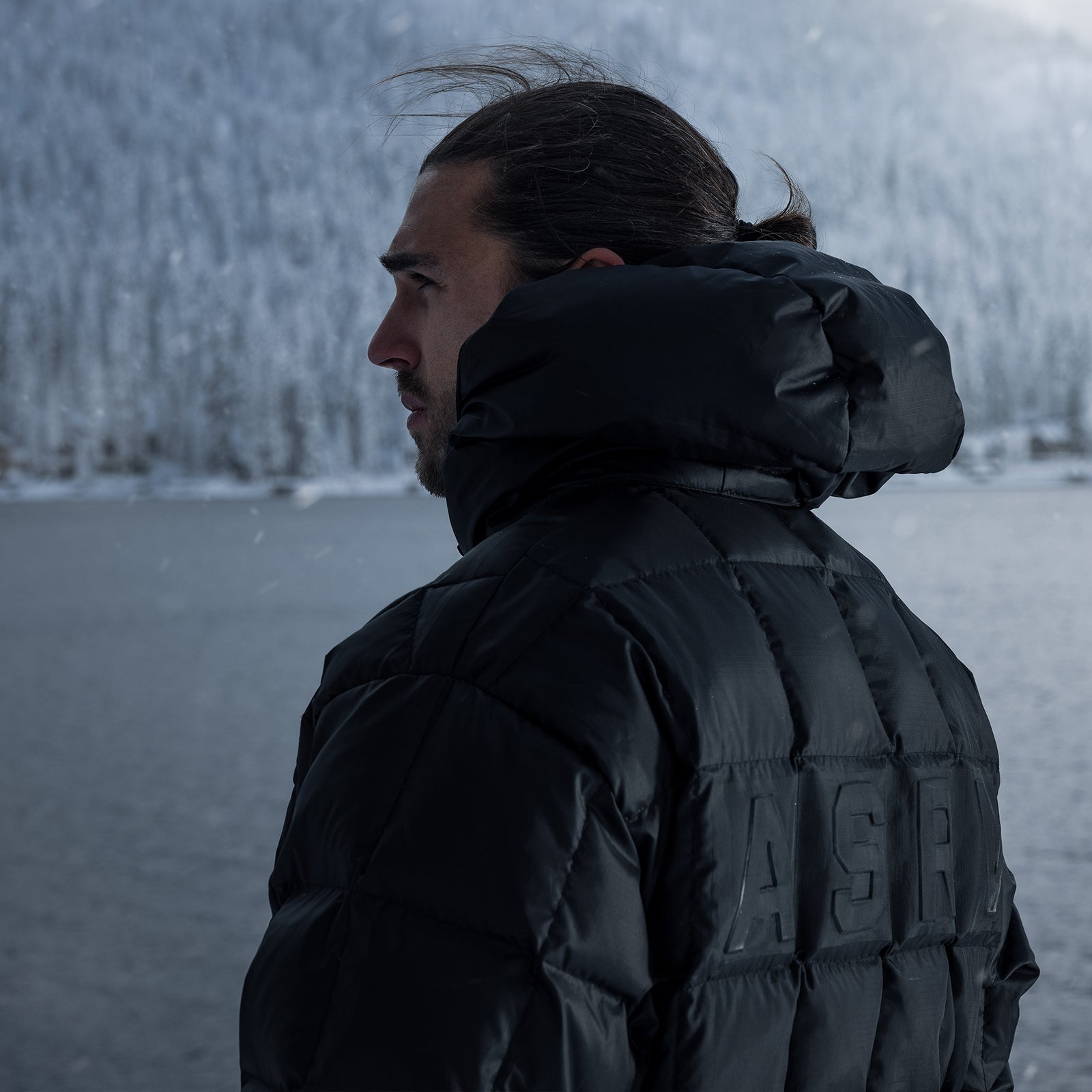 Black weather-ready down puffer jacket worn by model in profile, overlooking snowy landscape