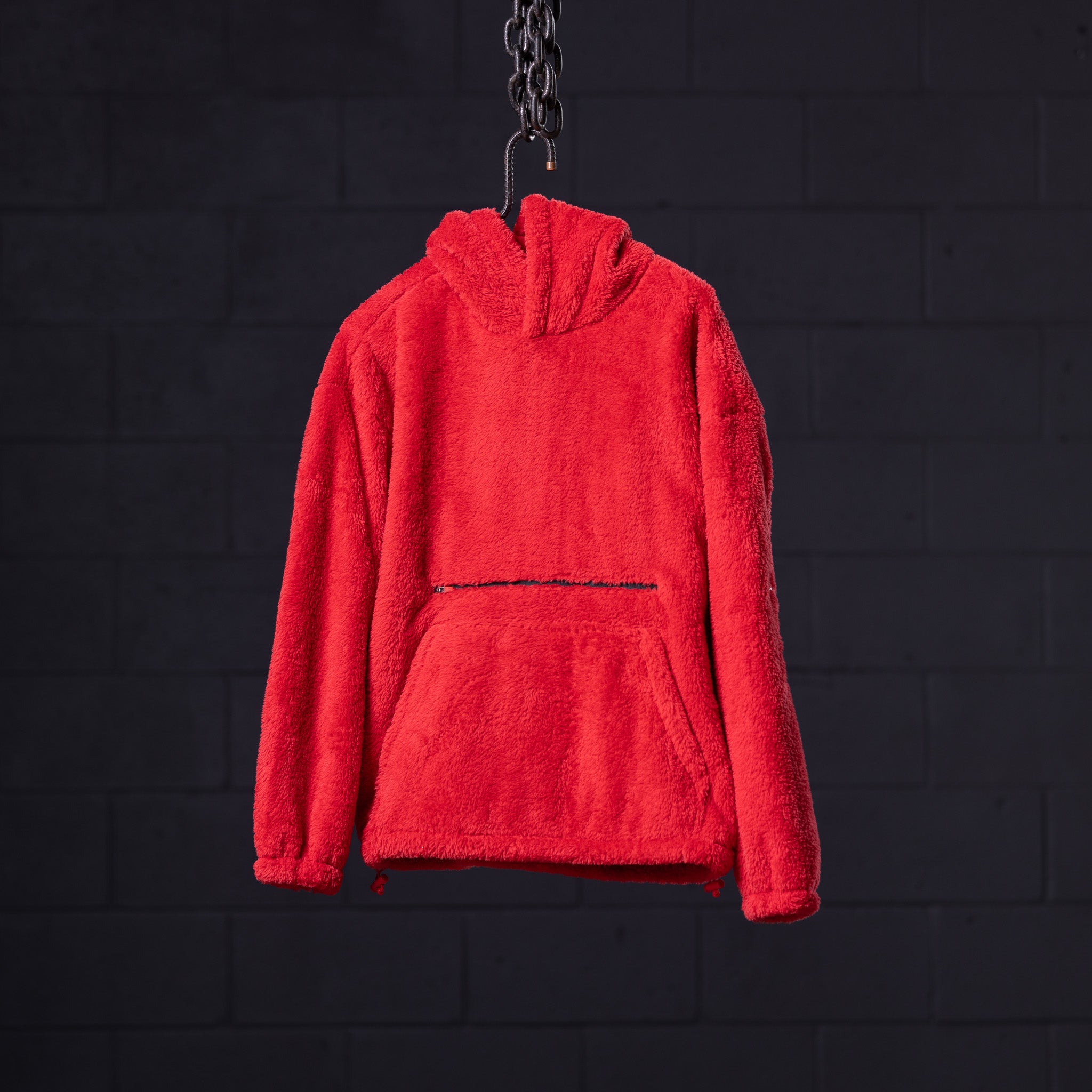 Red sherpa recovery hoodie hanging on chain against dark brick background