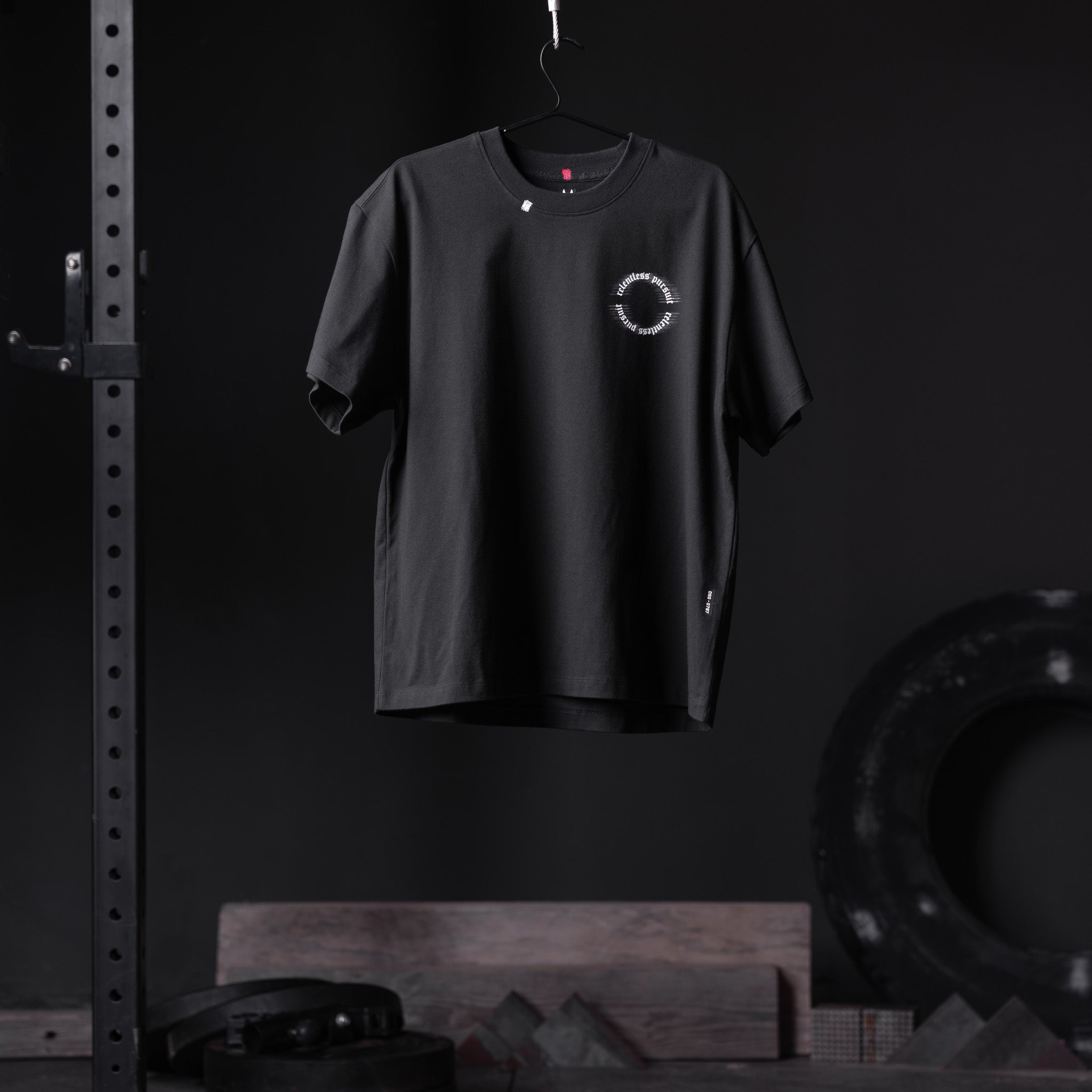 Black Tech Essential Relaxed Tee with circular logo, displayed on hanger in gym setting