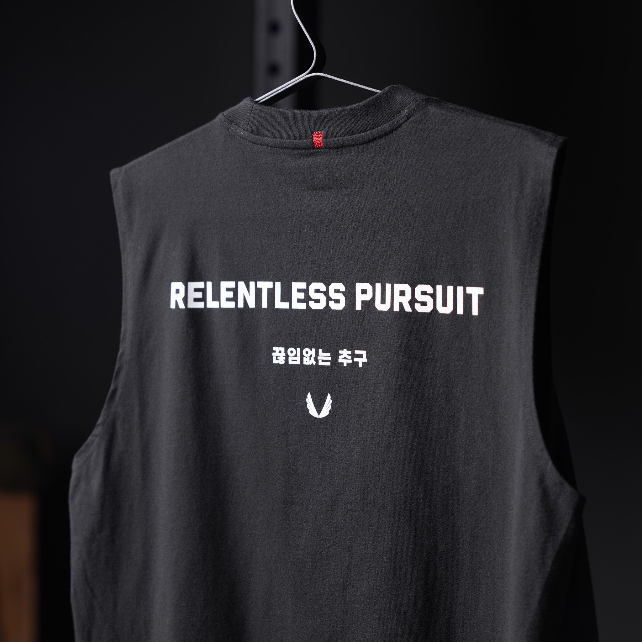 Black Tech Essential Relaxed Cutoff sleeveless tank on hanger, back view showing 'Relentless Pursuit' text