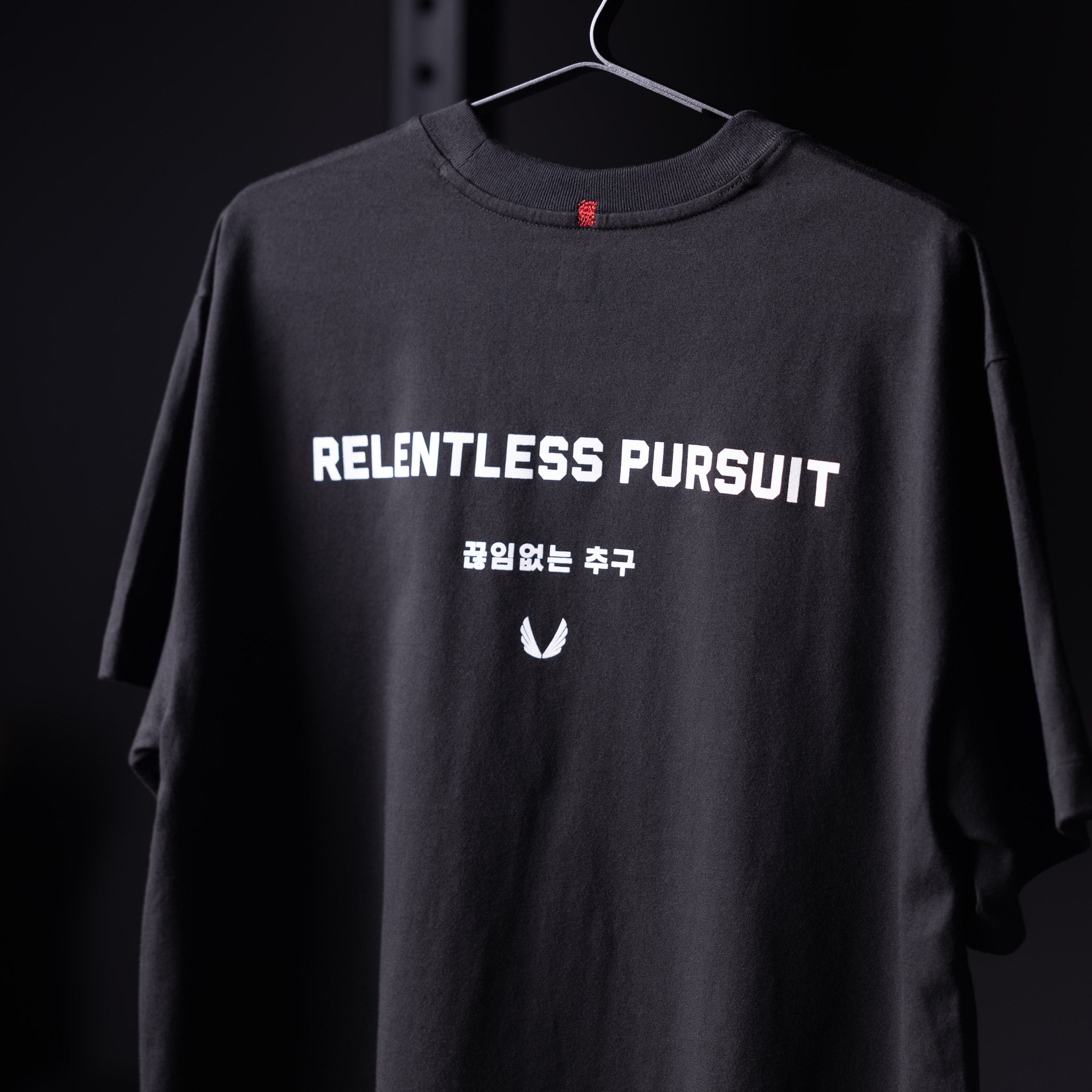 Black 'Relentless Pursuit' relaxed tee on hanger, front view showing graphic print and Korean text