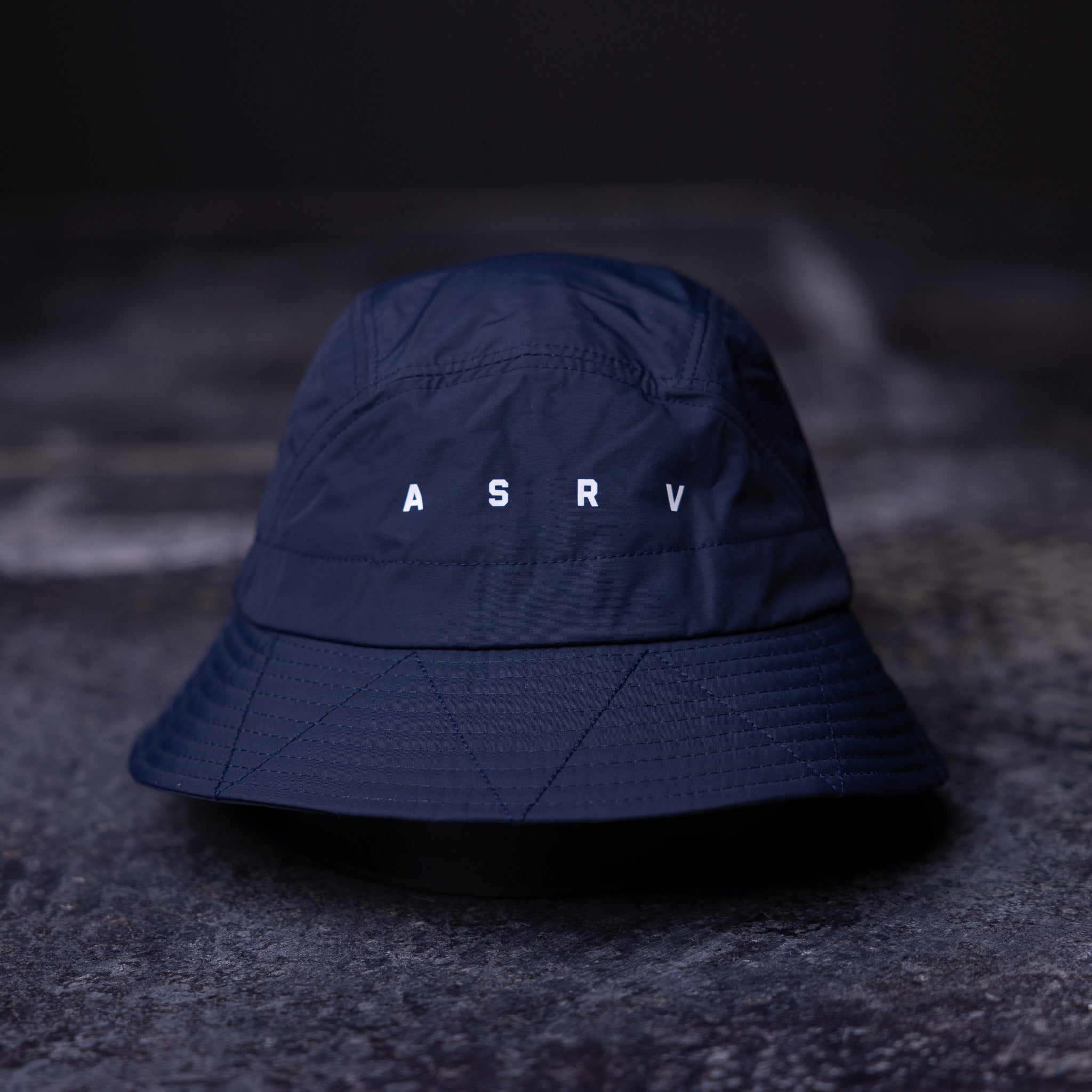 flatlay WN'22 Cinch Bucket Hat in navy blue, front view on dark surface