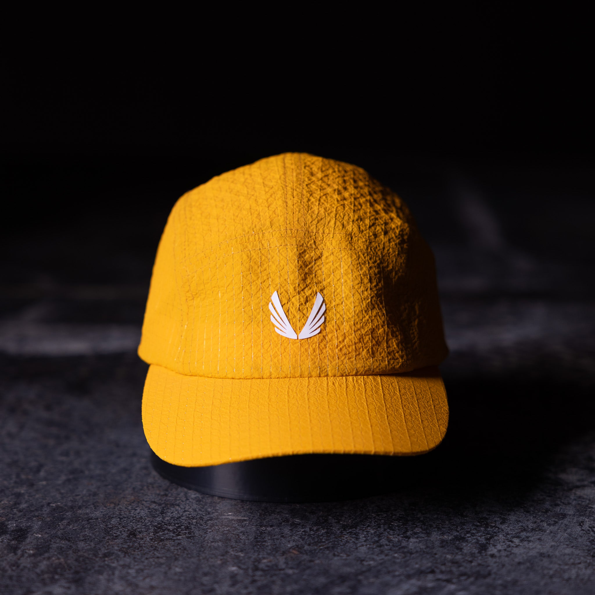 flatlay WN'23 5 Panel Hat in golden yellow, front view on dark surface