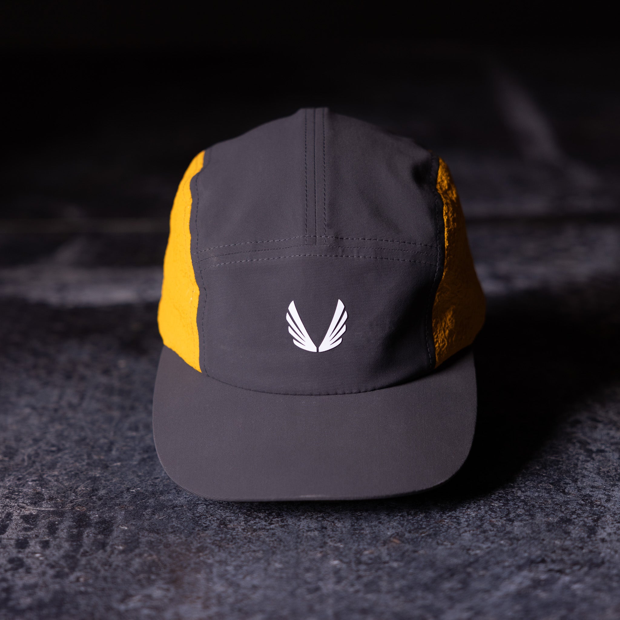 Flatlay of Two Tone 5 Panel Hat in charcoal and gold with white wing logo, front view
