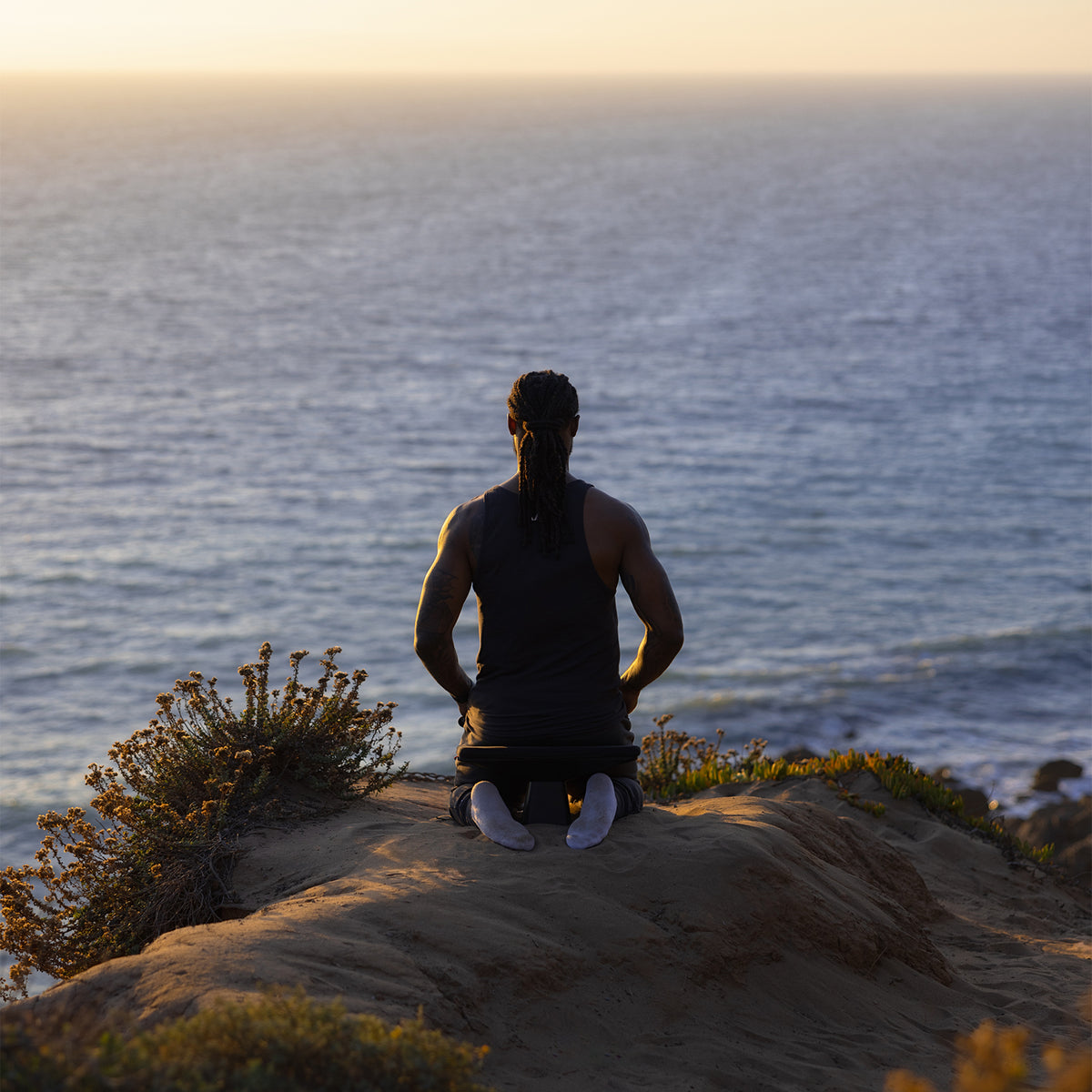 Man wearing black ASRV meditation bench tank top, seated on rocky cliff overlooking ocean at sunset