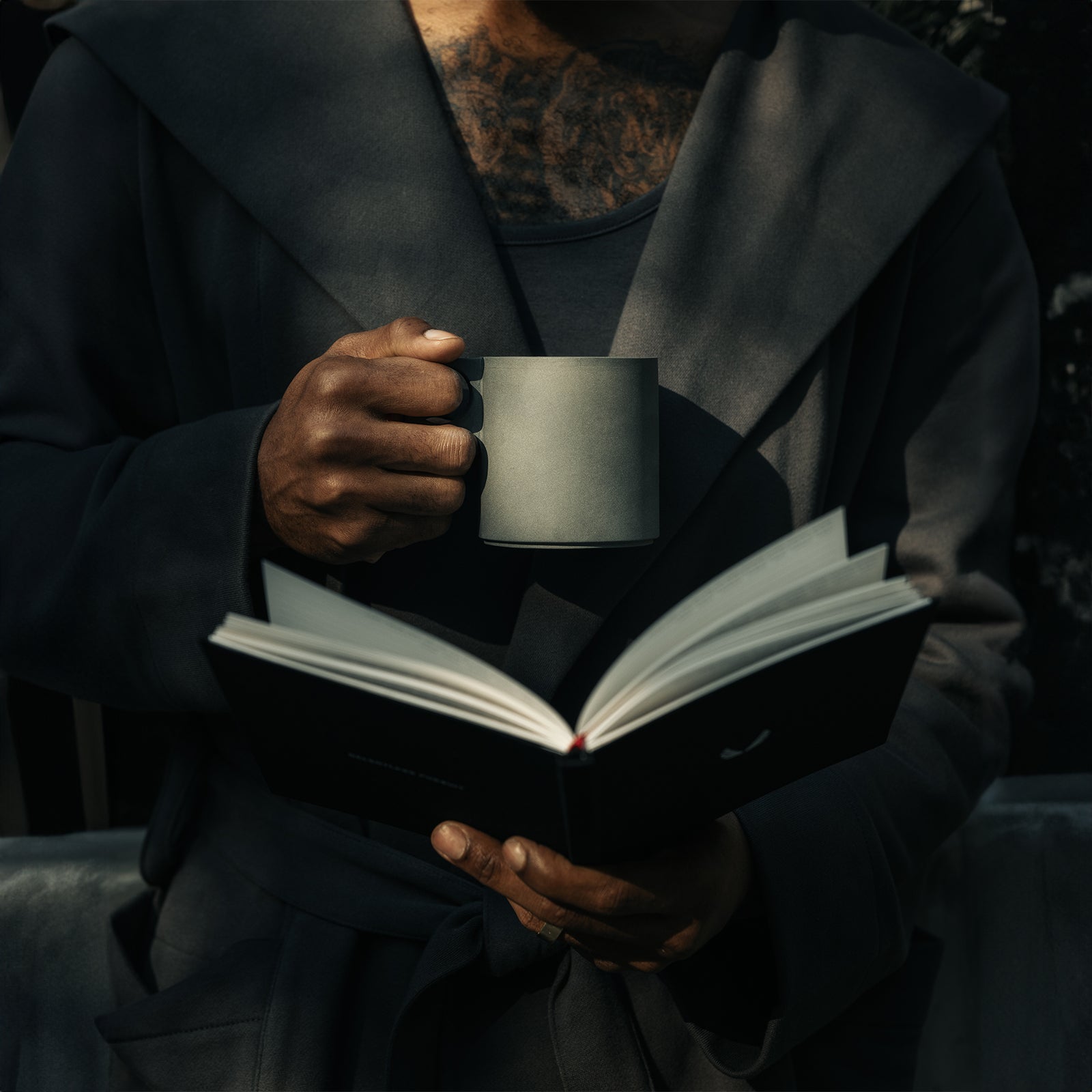Person in black hoodie holding white mug and open journal while reading